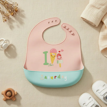 Saturday Ice Cream silicone baby bib flat lay showing soft blush pink bib with illustrated smiling ice cream cone and popsicle characters Ice Cream text with fresh mint teal food catch pocket stamped Saturday in multicolour lettering on linen background with wooden turtle teether white pacifier and white lace-up sneakers