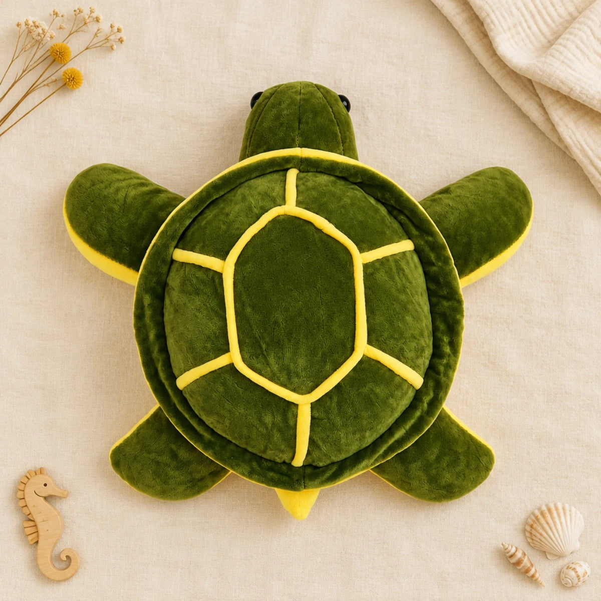 Giant green sea turtle plush toy flat lay from directly above on cream linen background showing full top-down view of domed forest green shell with vivid yellow hexagonal scute piping four wide flat green flippers with yellow edges long textured green neck and two glossy black bead eyes beside wooden seahorse toy white scallop shells and dried yellow flowers