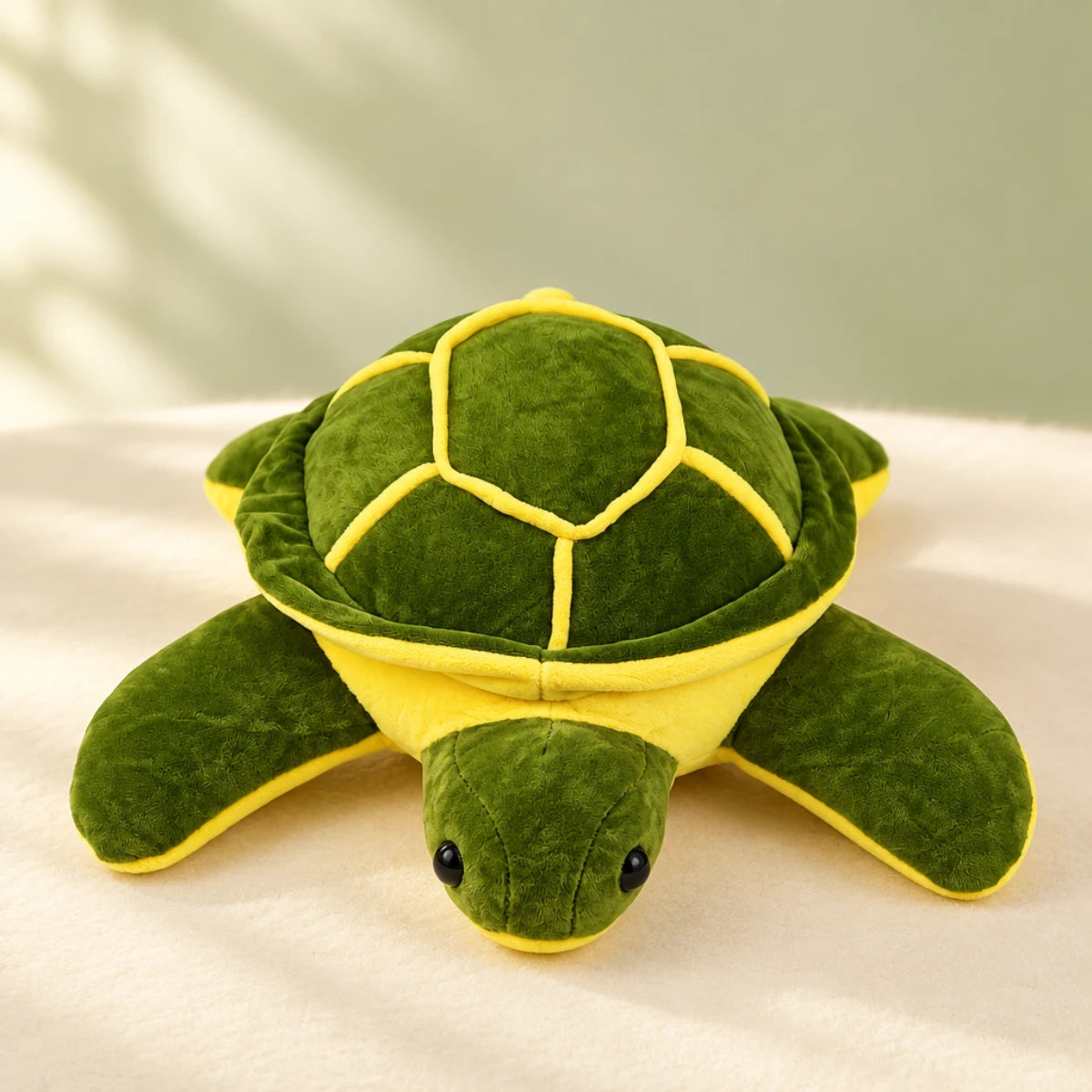 Giant green sea turtle plush toy studio scene lying flat on cream fabric surface with soft sage green background and dappled leaf shadow showing full front view of forest green plush sea turtle with beautifully domed hexagonal shell outlined in vivid yellow piping four wide flat flippers extended long neck and two glossy black bead eyes