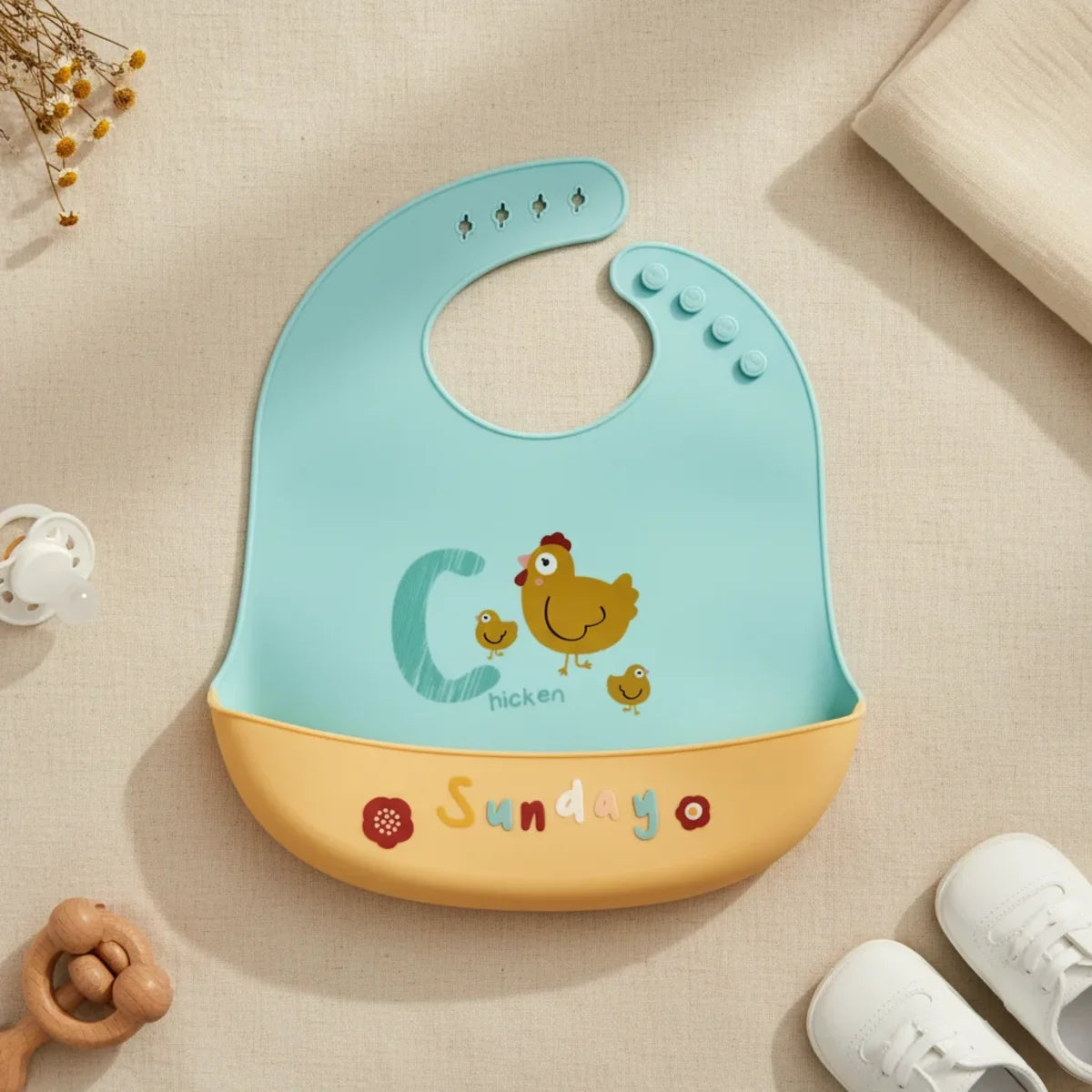 Sunday Chicken silicone baby bib flat lay showing fresh mint teal bib with illustrated mama hen and two baby chicks teal C letter and Chicken text with warm golden yellow food catch pocket stamped Sunday in multicolour lettering with red flower accent on linen background with wooden ring teether white pacifier and white lace-up sneakers