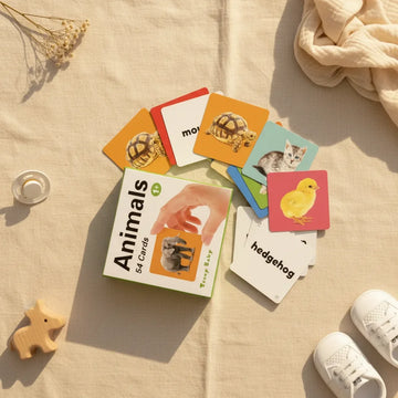 Teeny Baby Animals flash cards flat lay showing green Teeny Baby Animals 54 card gift box with photographic tortoise orange background kitten blue background chick pink background and white hedgehog word card fanned out on linen background with wooden elephant teether white pacifier and white baby sneakers