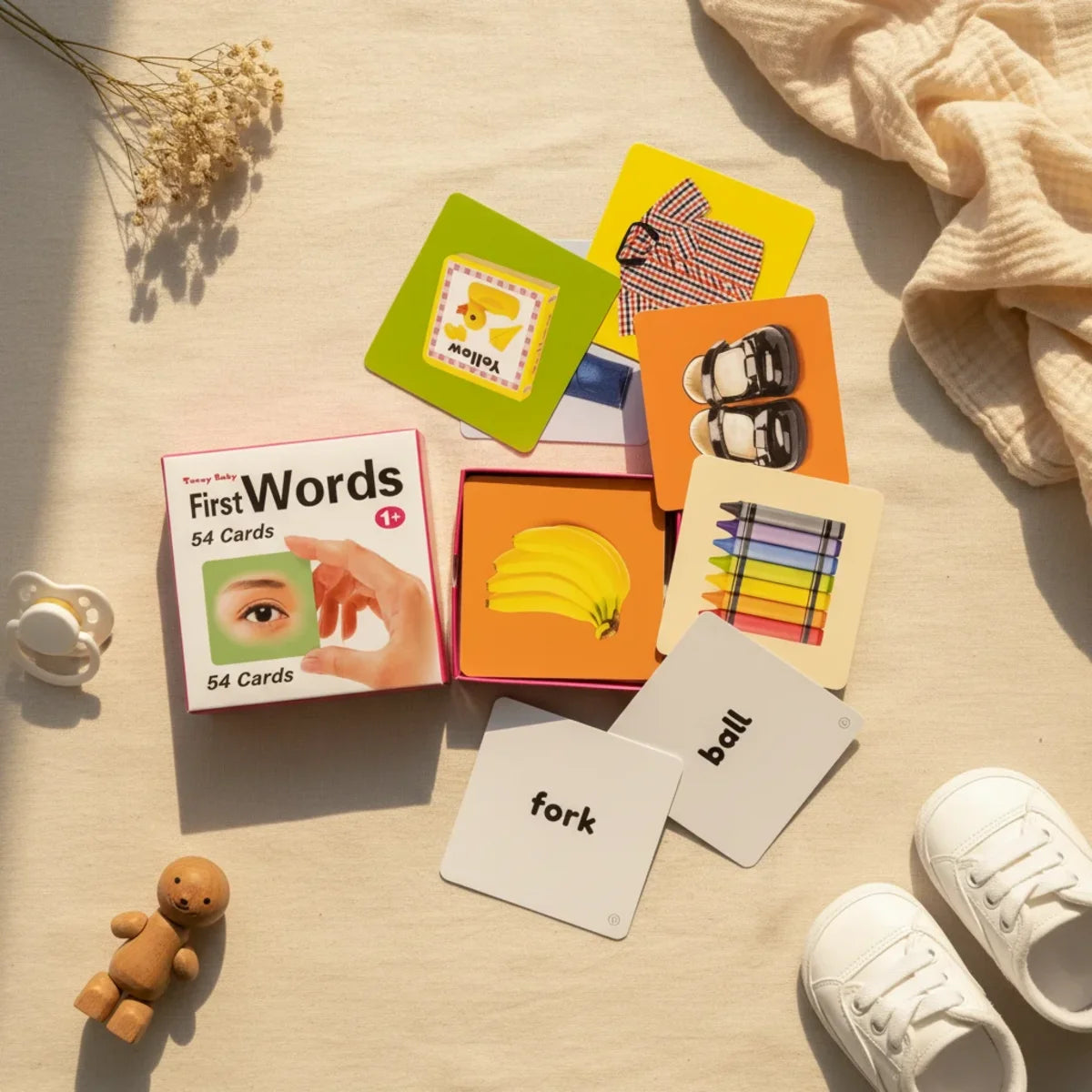 Teeny Baby First Words flash cards flat lay showing pink Teeny Baby First Words 54 card gift box with photographic banana orange background yellow duck green background shoes orange background crayons and white fork and ball word cards on linen background with wooden bear figurine white pacifier and white baby sneakers