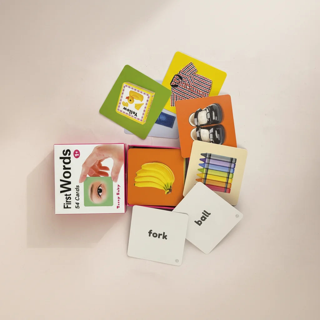Teeny Baby First Words flash cards studio flat lay showing pink Teeny Baby box with photographic banana orange background yellow duck green background shoes orange background crayons cream background cards and white fork and ball word cards on light pink background