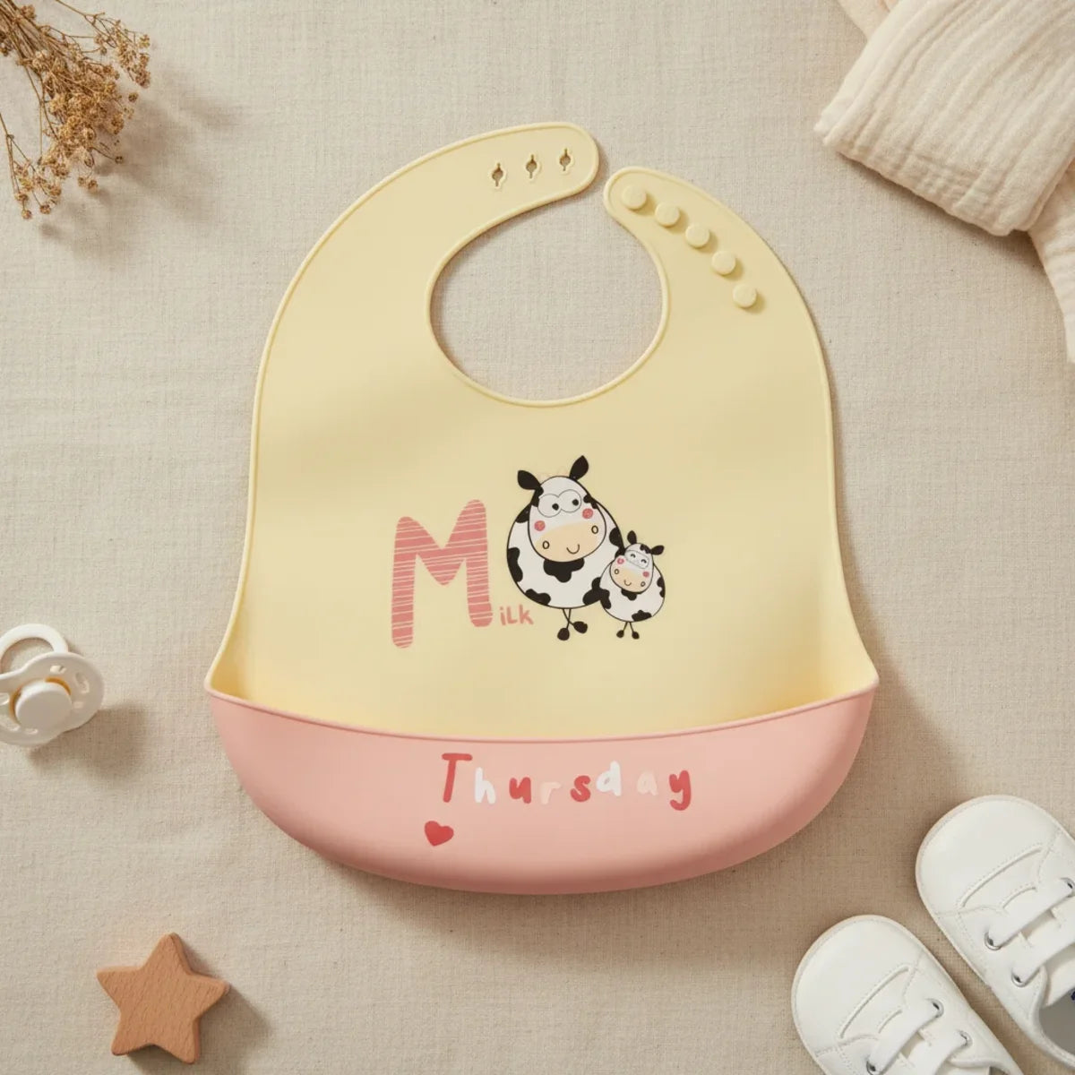 Thursday Milk Cow silicone baby bib flat lay showing soft butter yellow bib with illustrated chubby spotted mama and baby cow characters bold M letter and Milk text with blush pink food catch pocket stamped Thursday and red heart on linen background with wooden star toy white pacifier and white lace-up sneakers