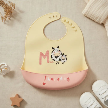 Thursday Milk Cow silicone baby bib flat lay showing soft butter yellow bib with illustrated chubby spotted mama and baby cow characters bold M letter and Milk text with blush pink food catch pocket stamped Thursday and red heart on linen background with wooden star toy white pacifier and white lace-up sneakers