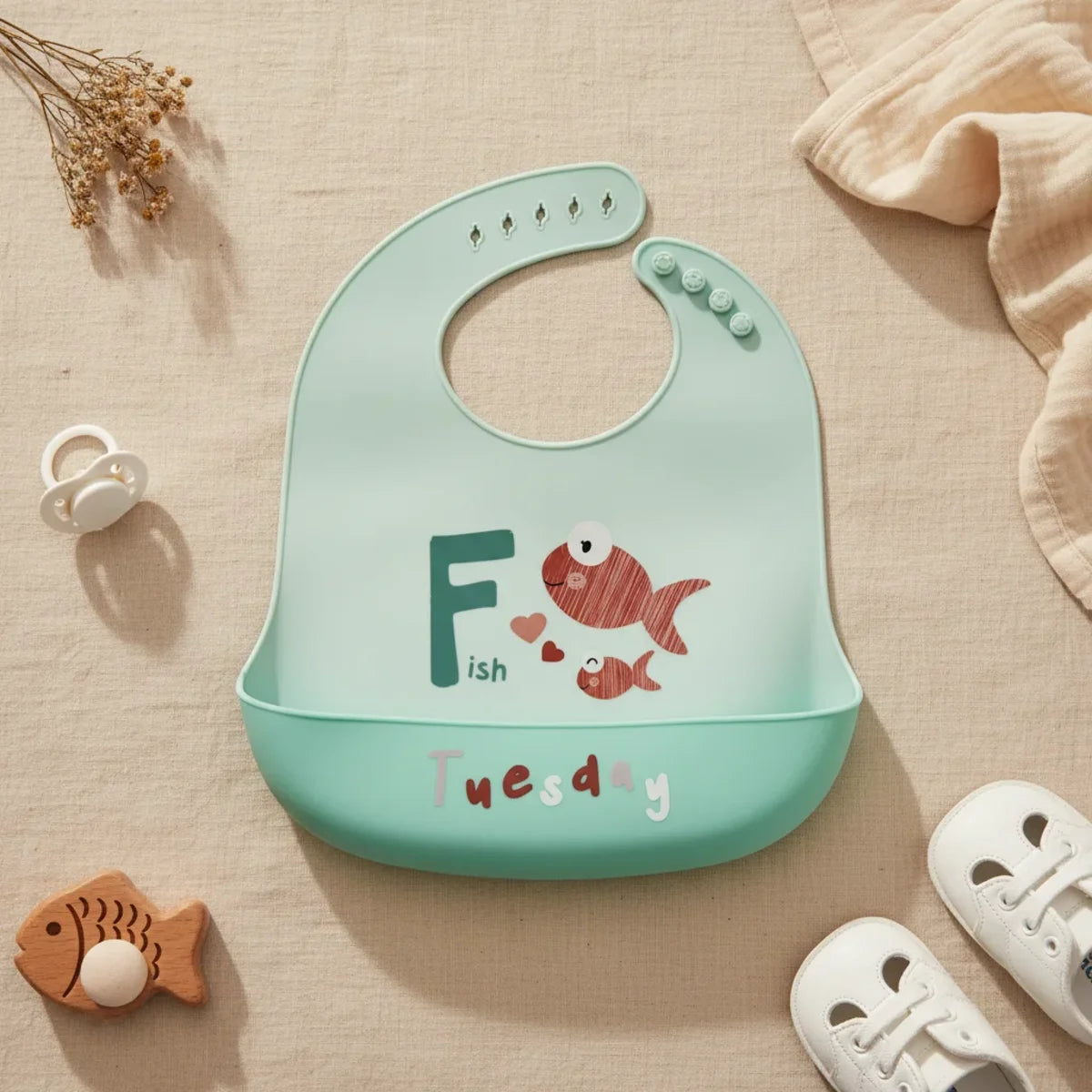 Tuesday Fish silicone baby bib flat lay showing all-mint teal bib with illustrated terracotta red mama and baby fish bold teal F letter Fish text and floating hearts with deeper teal food catch pocket stamped Tuesday in white and grey multicolour lettering on linen background with wooden fish teether white pacifier and white lace-up sneakers