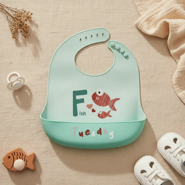 Tuesday Fish silicone baby bib flat lay showing all-mint teal bib with illustrated terracotta red mama and baby fish bold teal F letter Fish text and floating hearts with deeper teal food catch pocket stamped Tuesday in white and grey multicolour lettering on linen background with wooden fish teether white pacifier and white lace-up sneakers