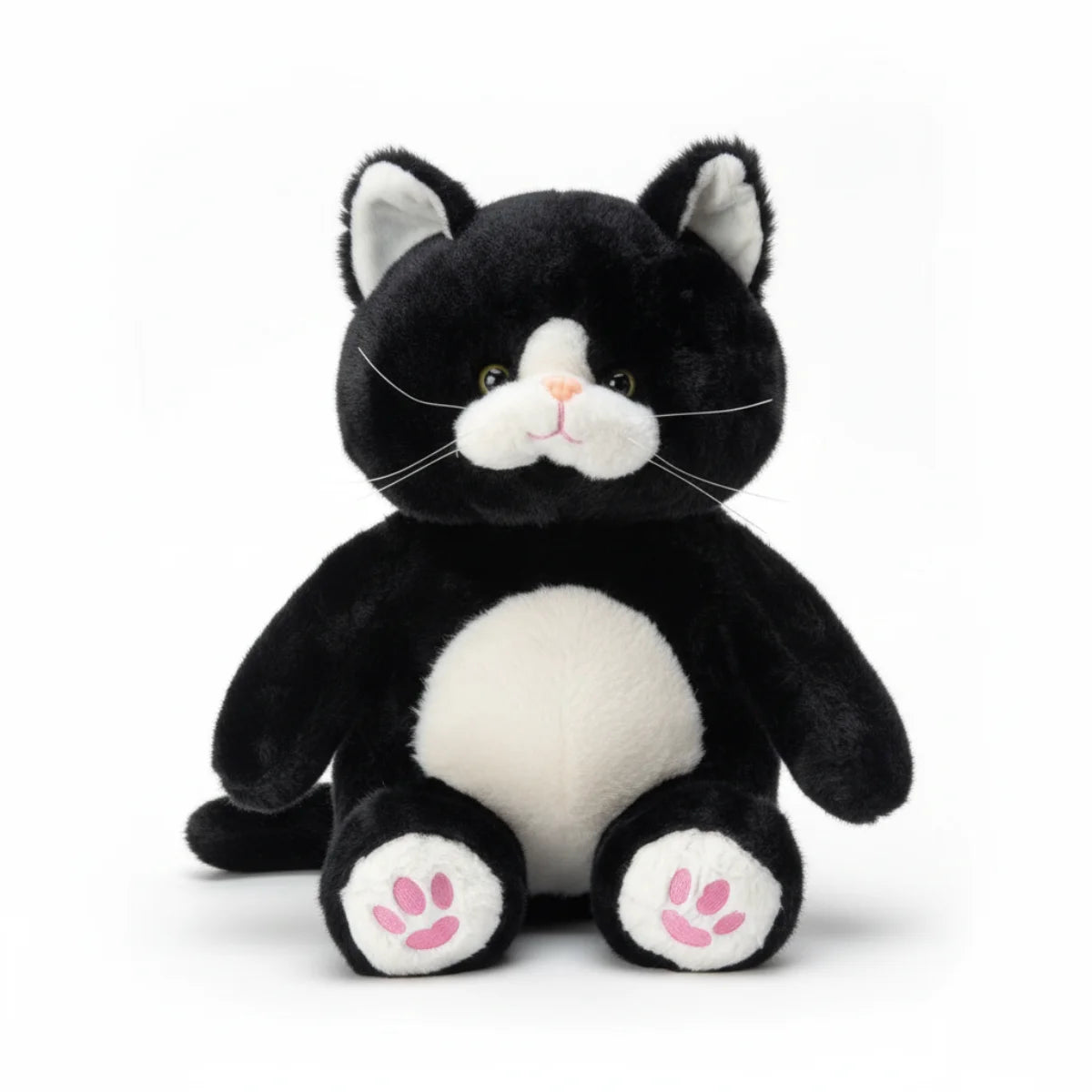 Tuxedo cat plush toy hero shot on white background showing full front face of chubby round black and white tuxedo kitten with vivid green eyes pink nose long silver thread whiskers fluffy white chest and belly white inner ears and oversized white feet with pink embroidered paw pad prints in sitting pose