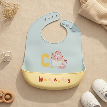 Wednesday Cupcake silicone baby bib flat lay showing soft periwinkle blue grey bib with illustrated smiling cupcake characters bold yellow C letter with soft yellow food catch pocket stamped Wednesday in red multicolour handwritten lettering on linen background with wooden ring teether white pacifier and white canvas sneakers