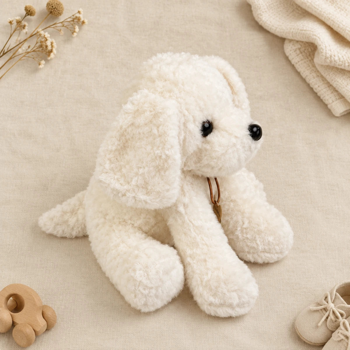 White fluffy puppy plush toy front face flat lay on cream linen background showing full front view of cloud-white curly bouclé Bichon style sitting puppy plush with long floppy ears two tiny glossy black bead eyes shiny black button nose gold ring chest detail and long front paws stretched forward in laid-back slouchy sitting pose beside cream knit throw dried flowers and white baby sneakers