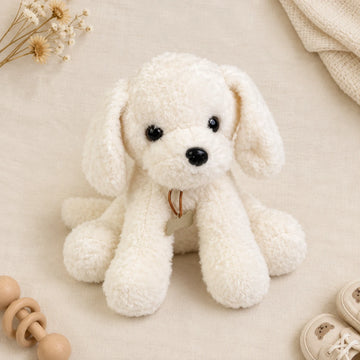 White fluffy puppy plush toy side angle flat lay on cream linen background showing three-quarter side view of cloud-white curly bouclé sitting puppy plush with long droopy floppy ears tiny glossy black bead eyes small black nose gold ring at chest and long front paws resting forward in relaxed slouchy pose beside wooden teether ring cream knit blanket and dried cotton stems