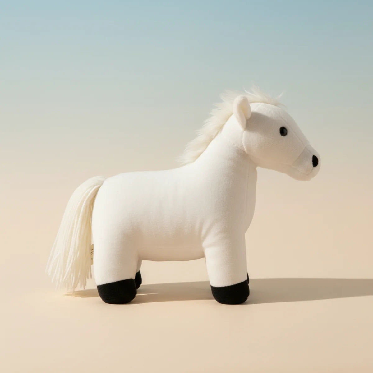 White horse plush toy studio scene on warm sand beige gradient background showing full side profile of snow white standing horse with flowing white mane cascading down neck full white tail bold black hooves glossy black button eyes and soft black nose in proud natural standing pose